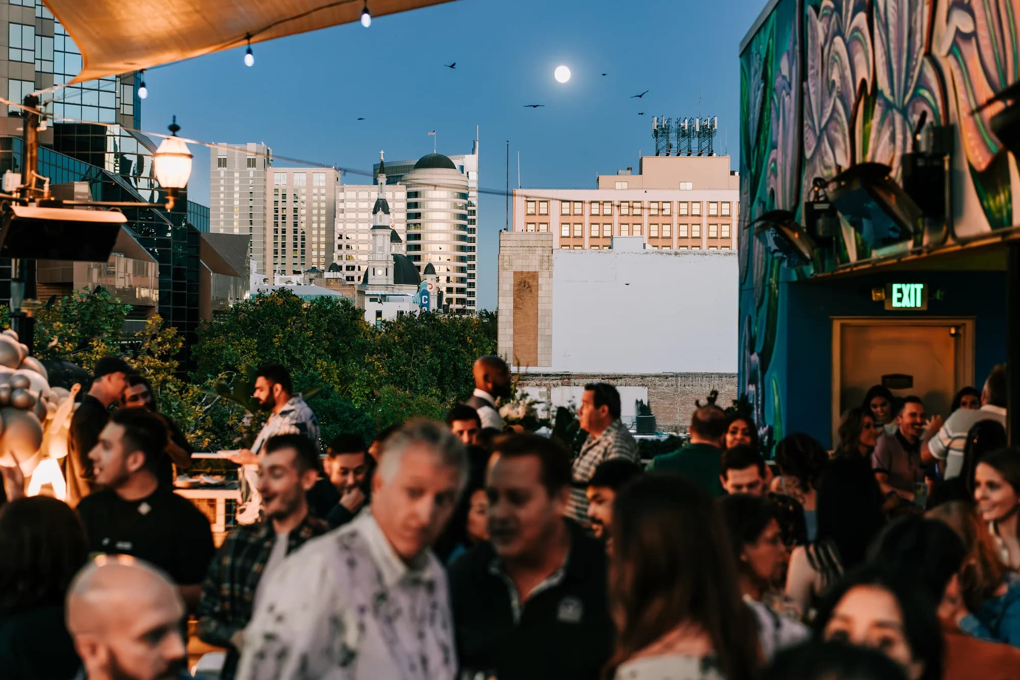 Darling Aviary rooftop buyout at night with a full moon over the Downtown Sacramento skyline, string lights and the Cathedral of the Blessed Sacrament dome visible from K Street