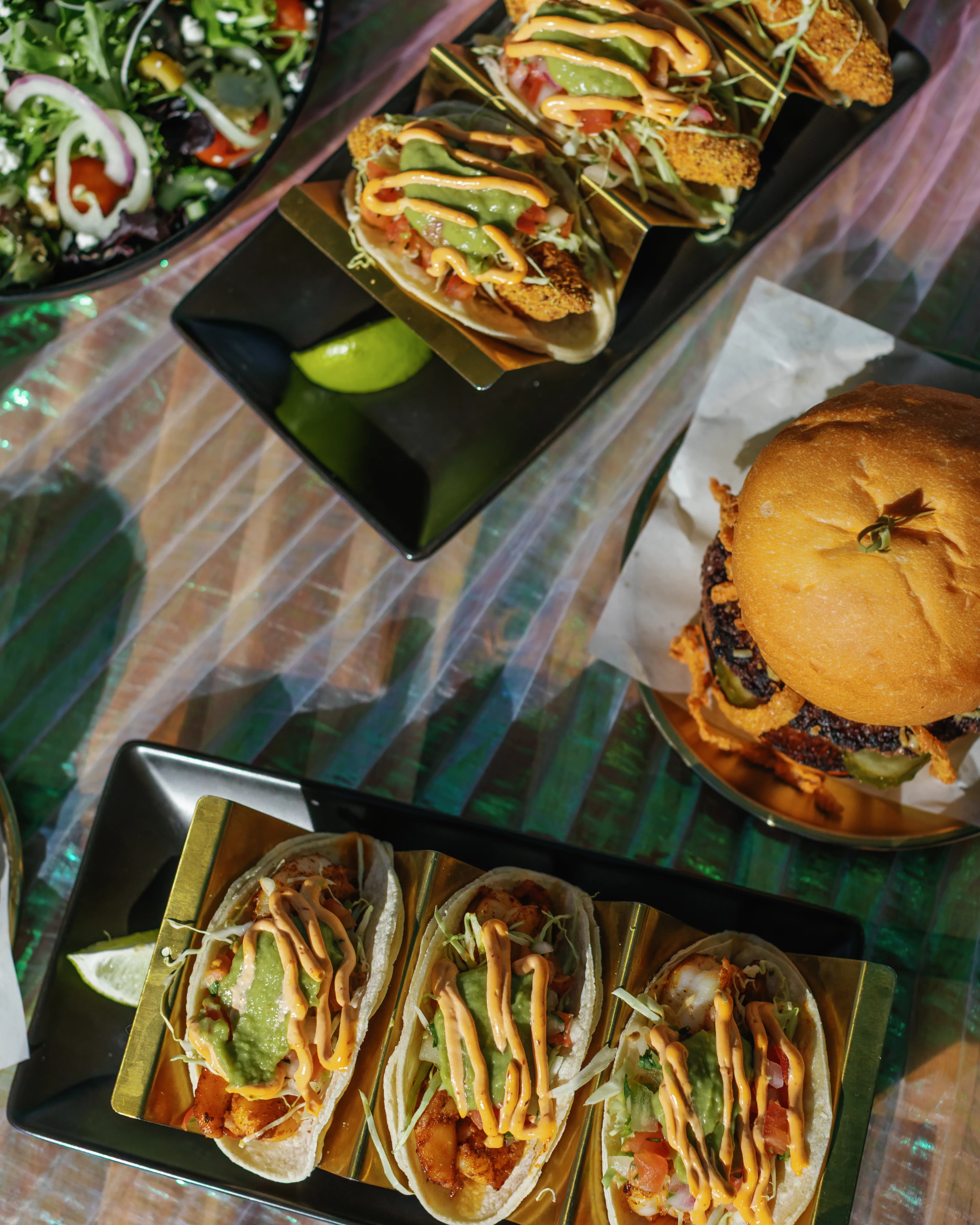 Overhead flatlay of Darling Aviary crispy fish tacos, crispy onion burger and side salad spread on an iridescent table — rooftop event menu at DoCo, Downtown Sacramento